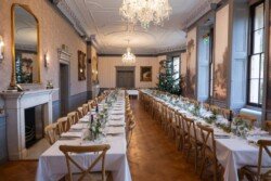 An elegant dining room at Hartham Park Weddings features long tables set for a meal, adorned with white tablecloths and floral arrangements. A Christmas tree stands gracefully at the far end, adding a festive touch to this enchanting setting.