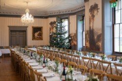 Elegant Hartham Park Weddings dining room set for an event, featuring long tables with white tablecloths, green floral arrangements, a decorated Christmas tree, and classic paintings on the walls.