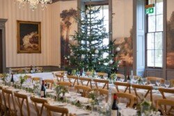An elegant dining room set for a banquet at Hartham Park Weddings, featuring long tables, wooden chairs, a beautifully decorated Christmas tree, and stunning paintings adorning the walls.