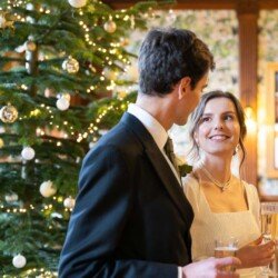 Evoto A couple in formal attire stands near a beautifully decorated Christmas tree at Hartham Park Weddings, holding glasses.