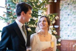 Evoto Elegantly dressed for the season, a couple stands before a beautifully decorated Christmas tree at Hartham Park Weddings, holding champagne glasses and sharing smiles full of festive cheer.
