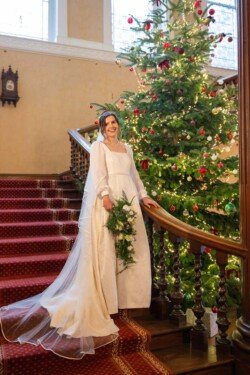 Evoto A bride in a white dress stands on a red-carpeted staircase at Hartham Park Weddings, next to a decorated Christmas tree, holding a bouquet of green and white flowers.
