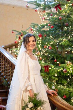 Evoto A bride stands by a decorated Christmas tree at Hartham Park Weddings, smiling. She wears a white dress and veil, holding a bouquet. The tree is adorned with red ornaments and lights, adding festive charm to the elegant setting.
