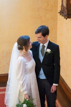 A bride and groom smile at each other in an ornate room at Hartham Park Weddings. The bride wears a white dress and veil, while the groom is in a black suit with a blue vest.