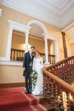 A couple in formal wedding attire stands on a wooden staircase in an ornate interior with arched doorways, embodying the timeless elegance of Hartham Park Weddings.