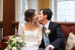 At Hartham Park Weddings, a bride and groom share a tender kiss while seated on a couch, the bride's hands gently cradling a bouquet of white flowers.