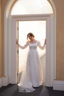 Evoto A woman in a long white dress stands in a doorway at Hartham Park Weddings, gently holding the doorframe with both hands.