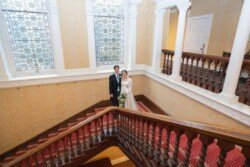 A couple stands on a grand staircase in the historic Hartham Park Weddings venue, with ornate stained glass windows in the background.