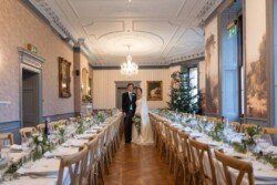 A bride and groom stand smiling in the elegant banquet hall of Hartham Park Weddings, surrounded by long, set tables and a beautifully adorned Christmas tree in the background.