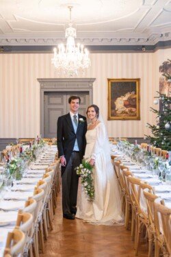 A couple stands together in the decorated banquet room at Hartham Park Weddings, with a long table set for an event. The elegant chandeliers and paintings on the walls add charm, while the bride clutches a floral bouquet.