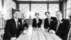 Five men in suits sit at a wooden table in a room with a patterned wall, reminiscent of the elegant charm found at Hartham Park Weddings.
