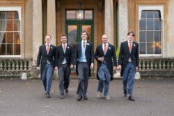 Five men in formal attire stride confidently towards the camera, the grandeur of columns behind them hinting at the elegance of a Hartham Park Weddings setting.