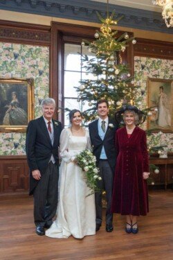 A bride and groom stand between two elegantly dressed guests in front of a decorated Christmas tree inside a room adorned with floral wallpaper and framed paintings, capturing the timeless elegance of Hartham Park Weddings.