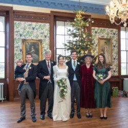 A wedding group photo at Hartham Park Weddings captures six adults and a child in elegant formal attire, standing before a beautifully decorated Christmas tree, surrounded by framed paintings in the charming venue.