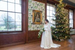 A bride in a white dress stands beside a decorated Christmas tree, the elegance of Hartham Park Weddings reflected in the room's large windows and patterned wallpaper.