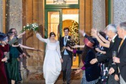 The bride and groom exit the building at Hartham Park Weddings, surrounded by cheering guests throwing confetti. The bride holds a bouquet and wears a stunning white dress, while the groom looks dapper in his suit and tie.