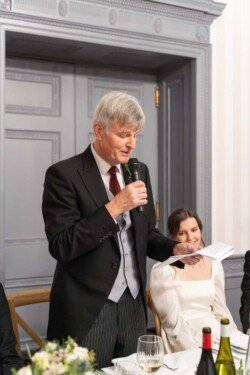 An elderly man in a suit speaks into a microphone while holding a paper, perhaps sharing heartfelt memories at Hartham Park Weddings. Nearby, a young woman sits smiling, adding warmth to the intimate gathering.
