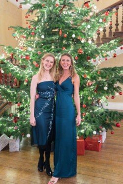 Two people wearing elegant blue dresses stand in front of a beautifully decorated Christmas tree at Hartham Park Weddings, with wrapped presents nestled beneath.