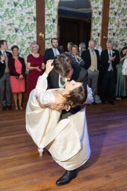 At a Hartham Park Wedding, a couple dances gracefully on a wooden floor, their elegance captivating the crowd. The woman in her flowing white dress and the man in his sharp suit share a magical moment as he gently dips her backward, surrounded by smiling onlookers.
