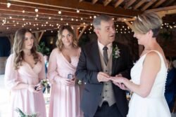 A man places a ring on a woman's finger during an Elmhay Park Weddings ceremony, while two women in pink dresses watch beside them under glowing string lights.