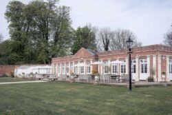 A long brick and glass building with outdoor seating and umbrellas sits beside a grassy lawn, bordered by tall trees and hedges—the perfect setting for Elmhay Park Weddings.