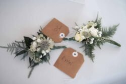 Two small floral boutonnieres with brown tags labeled "To" and "Ryan" rest on a white surface, perfect for elegant Elmhay Park Weddings.