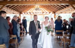 A bride and groom walk down the aisle in a wooden pavilion at Elmhay Park Weddings, smiling as guests stand and clap on either side.