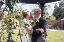 A couple dressed in formal attire stands outdoors under a decorative structure at Elmhay Park Weddings, smiling at each other and holding hands, surrounded by lush greenery.