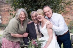 Two couples smile and pose outdoors at Elmhay Park Weddings; one woman in a white dress holds a bouquet, suggesting a wedding celebration. A brick wall and greenery create a charming backdrop.