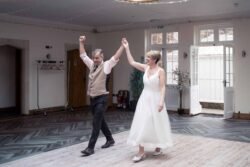 A man and woman, dressed formally, hold hands and smile while entering an empty room with wooden floors and white walls, capturing the elegance often seen at Elmhay Park Weddings.