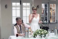 A woman in a white dress stands and speaks while holding a paper, next to a seated man at a beautifully decorated Elmhay Park Weddings table.