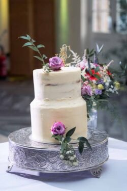 Two-tier white frosted cake decorated with purple roses and greenery, displayed on a silver stand—perfect for Elmhay Park Weddings—with a stunning floral arrangement in the background.
