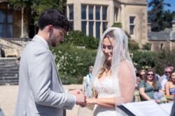 A groom places a ring on the bride's finger during an Orchardleigh House Weddings outdoor ceremony, with guests seated in the background.