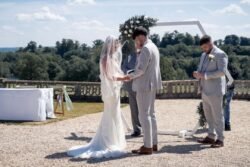 A bride and groom stand facing each other during an Orchardleigh House Weddings ceremony, with an officiant and groomsman nearby, under a beautifully decorated arch.