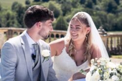 A bride and groom sit together outdoors at Orchardleigh House Weddings, smiling at each other. The bride wears a white dress and veil, while the groom is in a light gray suit with a white rose boutonnière.