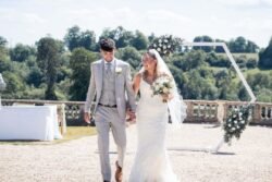 A bride in a white gown and a groom in a light grey suit walk hand in hand outdoors at Orchardleigh House Weddings, smiling, with floral decorations and greenery in the background.