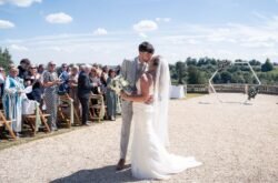 A bride and groom kiss outdoors at their Orchardleigh House Weddings ceremony, surrounded by guests seated in rows, with a geometric arch and scenic landscape in the background.