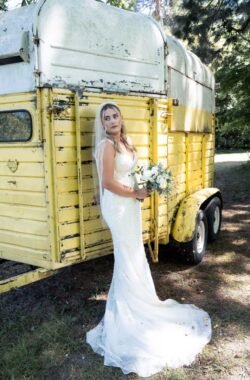 A bride in a white gown and veil holding a bouquet stands beside a weathered yellow trailer, capturing the rustic charm often seen at Orchardleigh House Weddings.