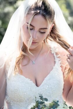 A bride in a white lace dress and veil looks down, holding part of her hair. She wears a necklace with an "A" pendant and stands outdoors, capturing the romantic charm of Orchardleigh House Weddings.