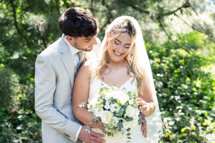 A bride in a white dress and veil holds a bouquet of white flowers while a groom in a light suit stands closely behind her outdoors at Orchardleigh House Weddings, both smiling.
