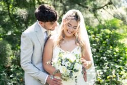 A bride in a white dress and veil holds a bouquet of white flowers while a groom in a light suit stands closely behind her outdoors at Orchardleigh House Weddings, both smiling.