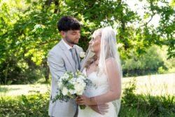 A bride in a white dress and veil holds a bouquet of white flowers while standing with a groom in a light gray suit outdoors, surrounded by greenery—capturing the elegance of Orchardleigh House Weddings.