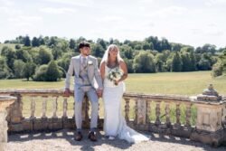 A bride and groom sit holding hands on a stone terrace at Orchardleigh House Weddings, dressed in wedding attire, with a green landscape and trees in the background.