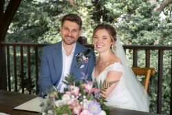 A bride and groom sit together at a table, smiling at the camera, with a bouquet of flowers in the foreground and lush greenery in the background—capturing the romantic charm of Haselbury Mill Weddings.