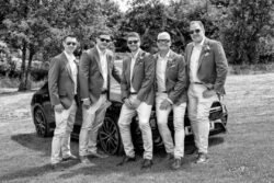 Five men in matching suits and sunglasses pose in front of a car on a grassy area with trees in the background, capturing classic style perfect for Haselbury Mill Weddings.