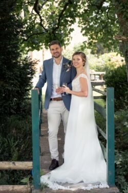 A man in a blue suit jacket and a woman in a white wedding dress stand together on a small bridge at Haselbury Mill Weddings, holding drinks and smiling at the camera, surrounded by lush greenery.