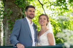 A bride and groom stand together outdoors at Haselbury Mill Weddings, dressed in wedding attire, smiling at the camera with green foliage in the background.