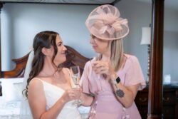 Two women dressed for a formal occasion stand indoors at a Bath Spa Hotel Weddings event, facing each other and smiling while holding glasses of champagne. One wears a bridal gown, the other a pink dress with a matching fascinator.