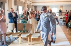 Two men in gray suits and sunglasses walk down the aisle at a Bath Spa Hotel Weddings ceremony as guests stand, smile, and applaud.
