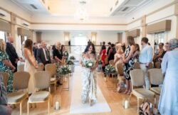 A woman in a floral dress walks down an aisle holding a bouquet, while guests seated on both sides watch in a brightly lit room with chandeliers at a stunning Bath Spa Hotel Weddings venue.